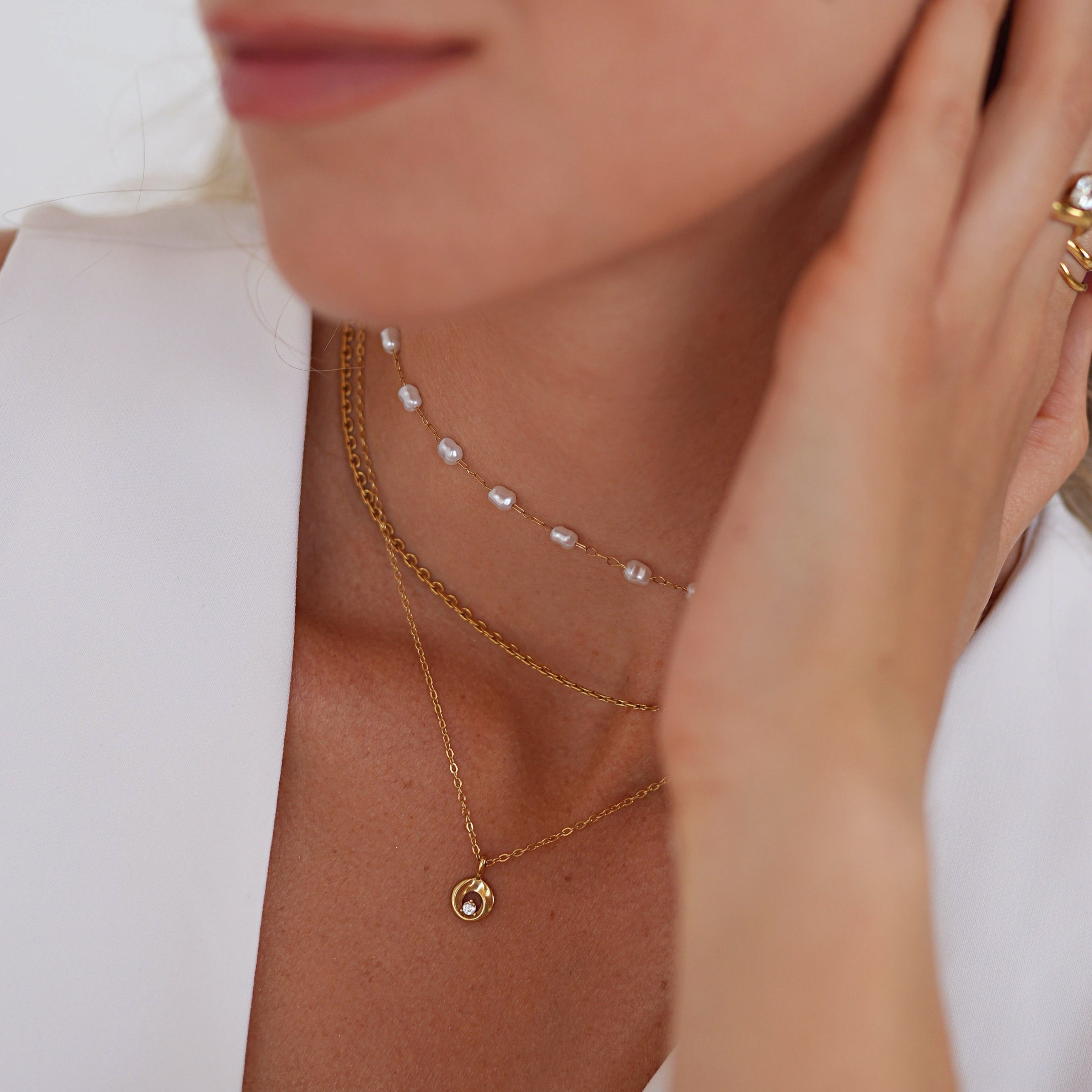 A close-up lifestyle shot of a woman wearing delicate gold jewelry, including layered necklaces, a ring, and a stud earring, paired with a white sleeveless top.