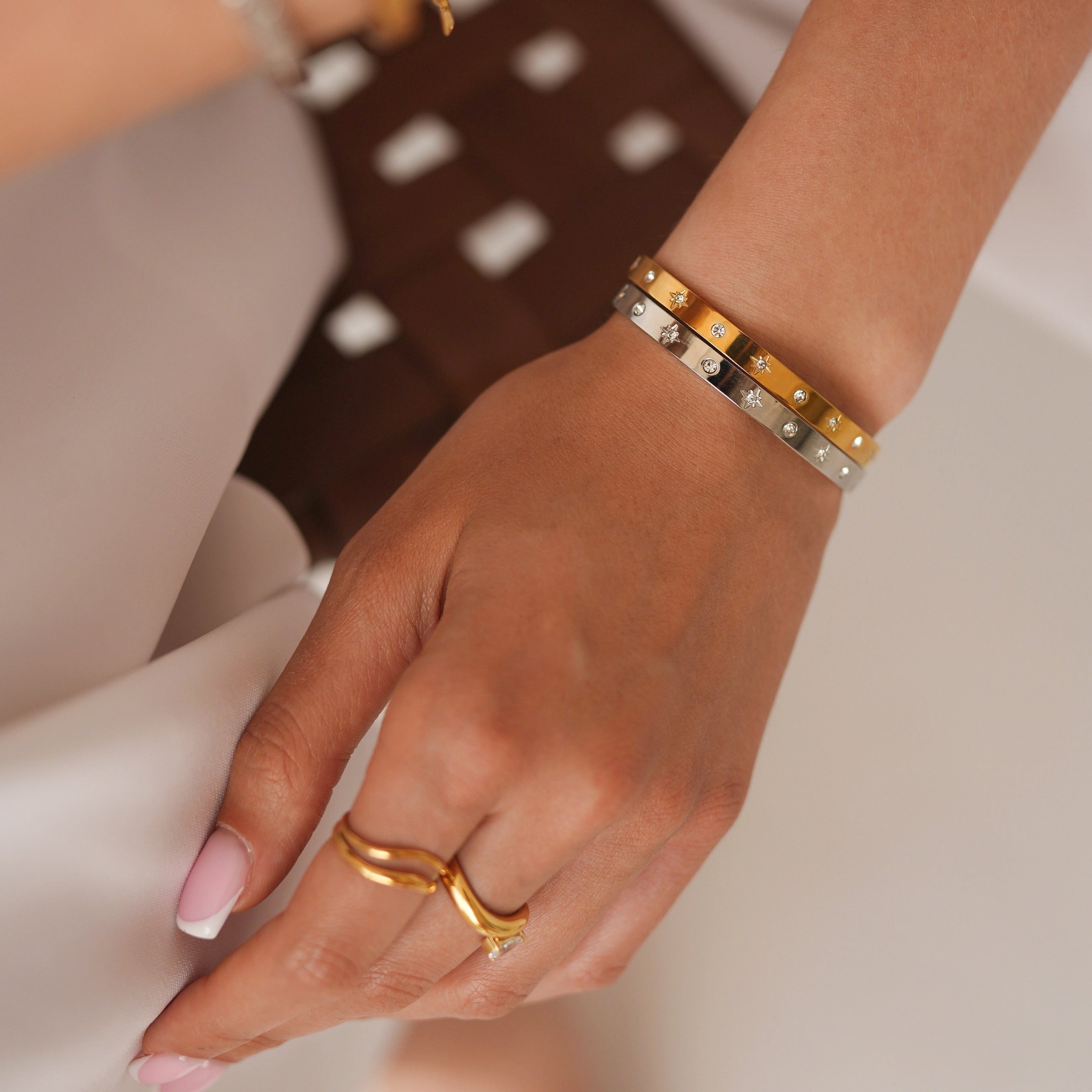A close-up of a person's hands and wrists wearing multiple bracelets and rings. The most prominent product is a gold bangle bracelet with alternating gold and silver-toned square links, worn on the wrist.