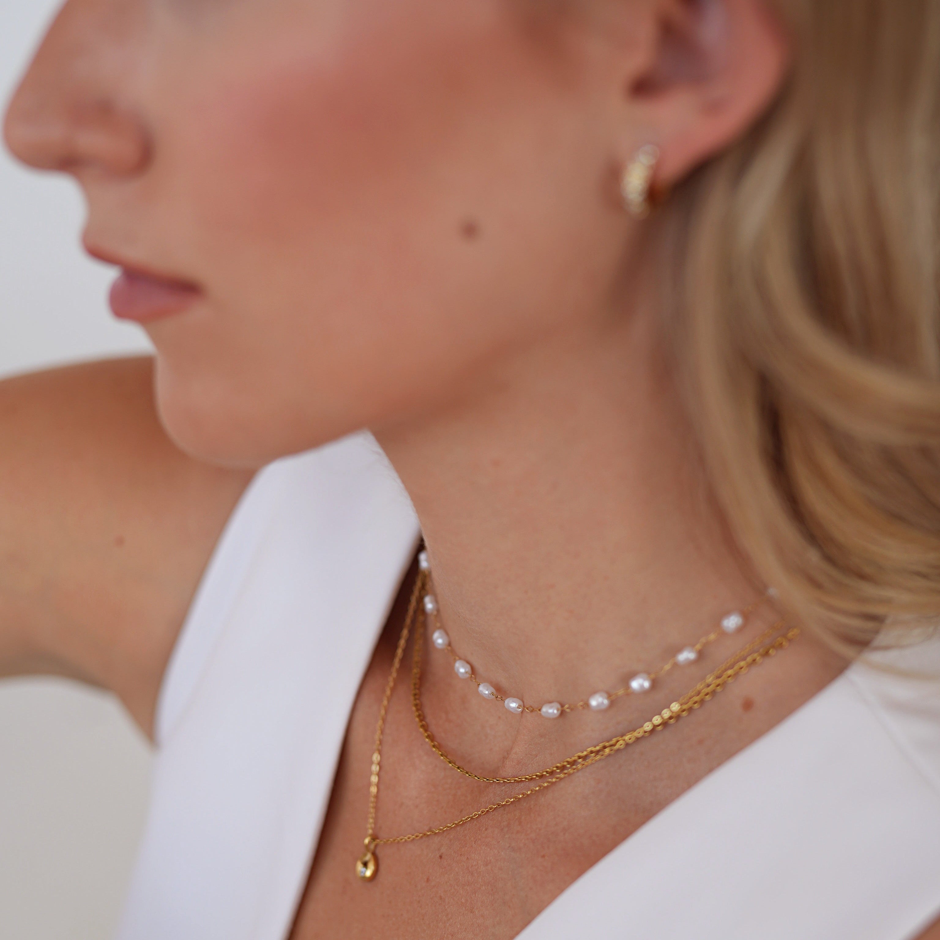 Close-up of a woman wearing layered necklaces, including a gold chain with a small teardrop pendant, a delicate gold chain, and a pearl necklace, paired with a white sleeveless top.