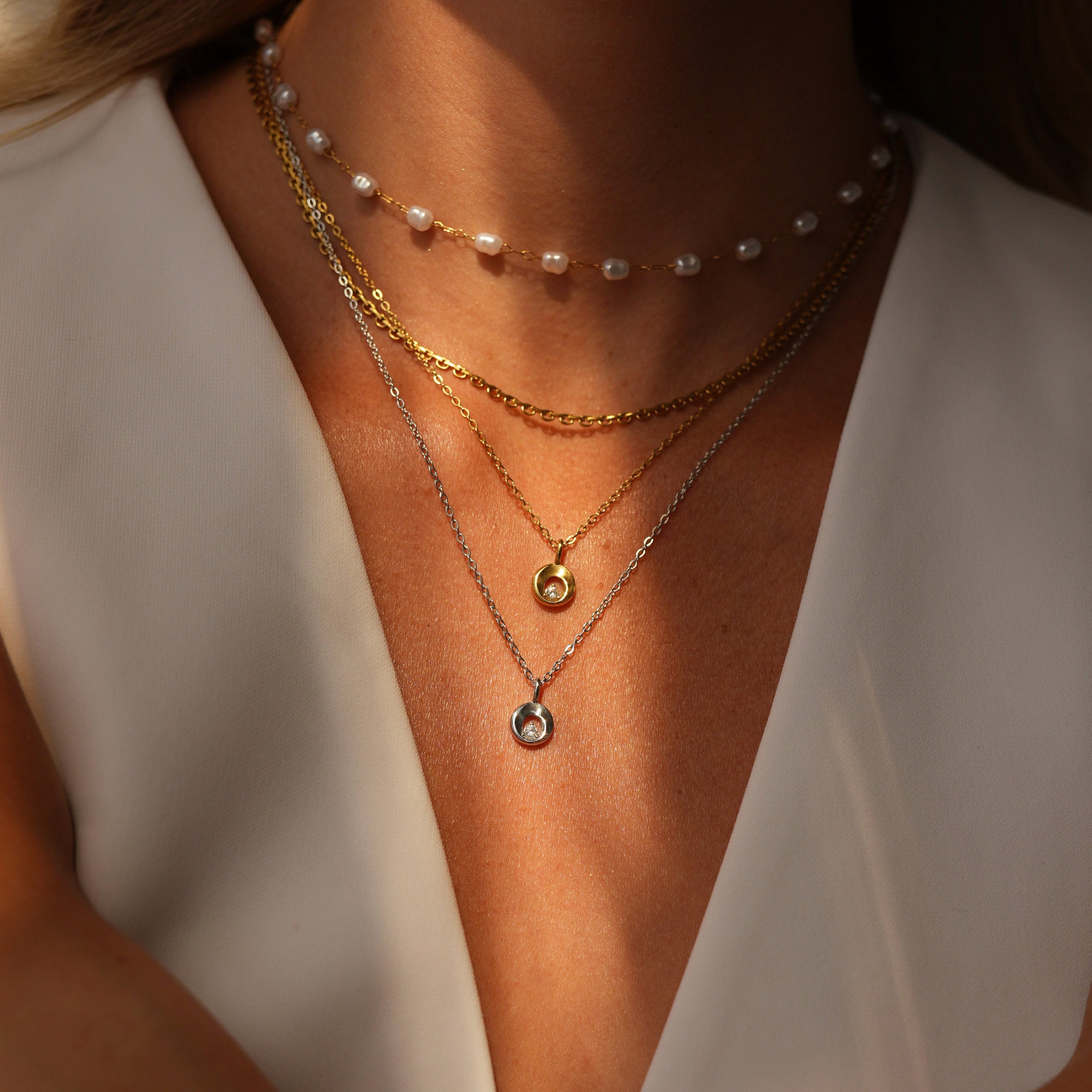 A close-up of a model wearing layered gold necklaces, including a pearl choker, a delicate chain, and a teardrop pendant necklace, paired with a white V-neck top.