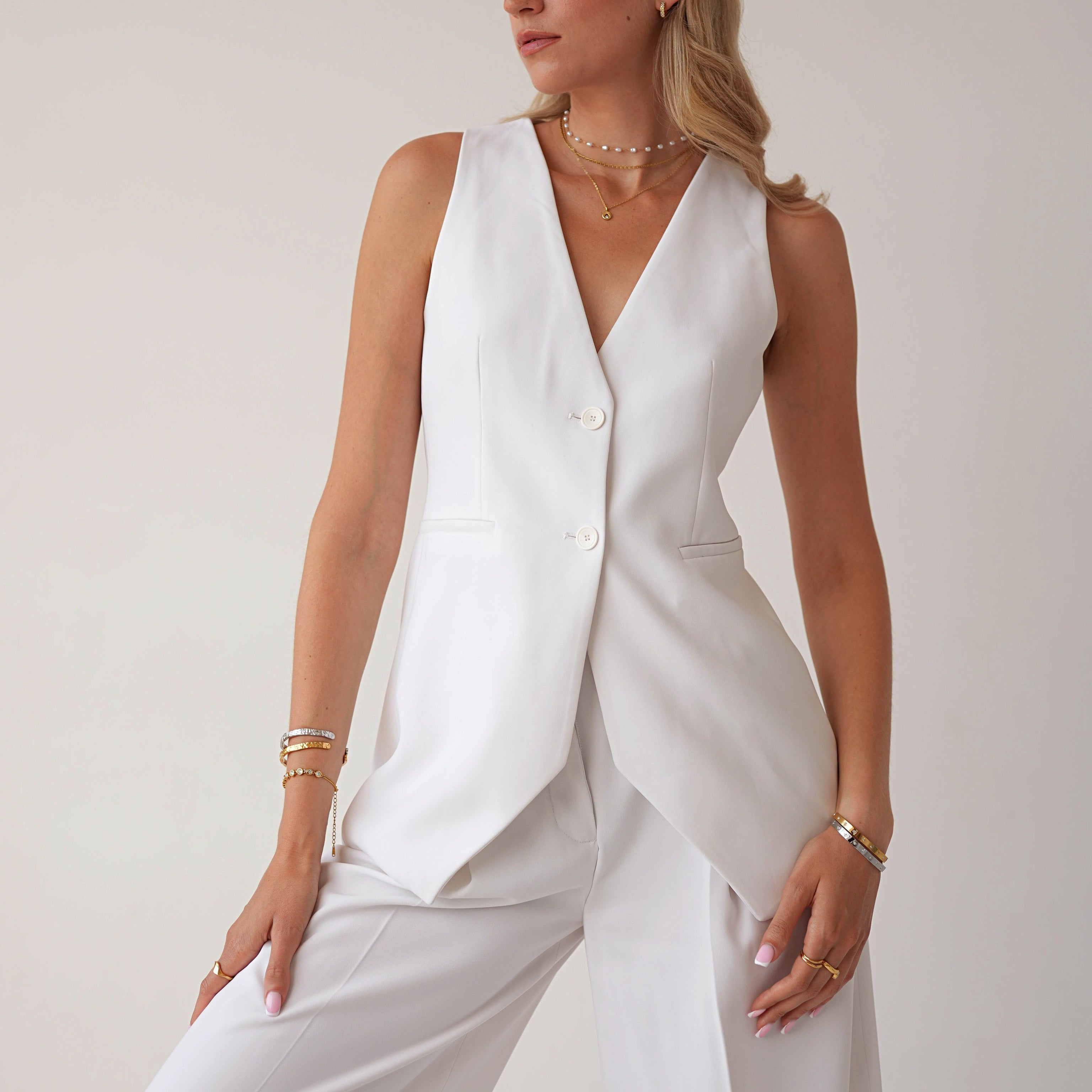 A woman modeling a white sleeveless blazer vest with a V-neck and front buttons, paired with matching white wide-leg trousers, against a plain light beige studio background.
