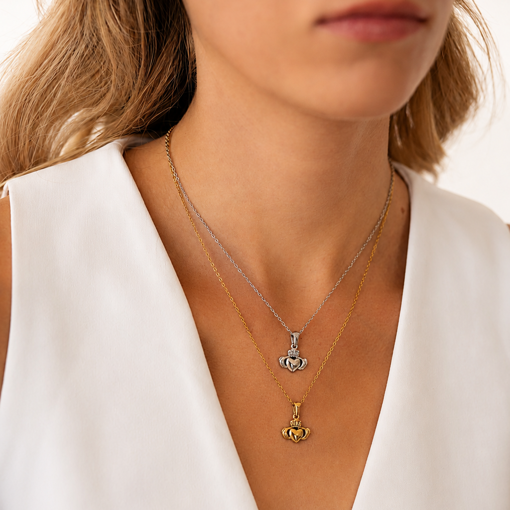 Model wearing two layered Claddagh necklaces — one silver, one gold — on a delicate chain. Both pendants feature the classic Claddagh design: crown, heart, and hands. Styled with a white sleeveless top. Shot against a warm neutral background. Available in silver and gold.