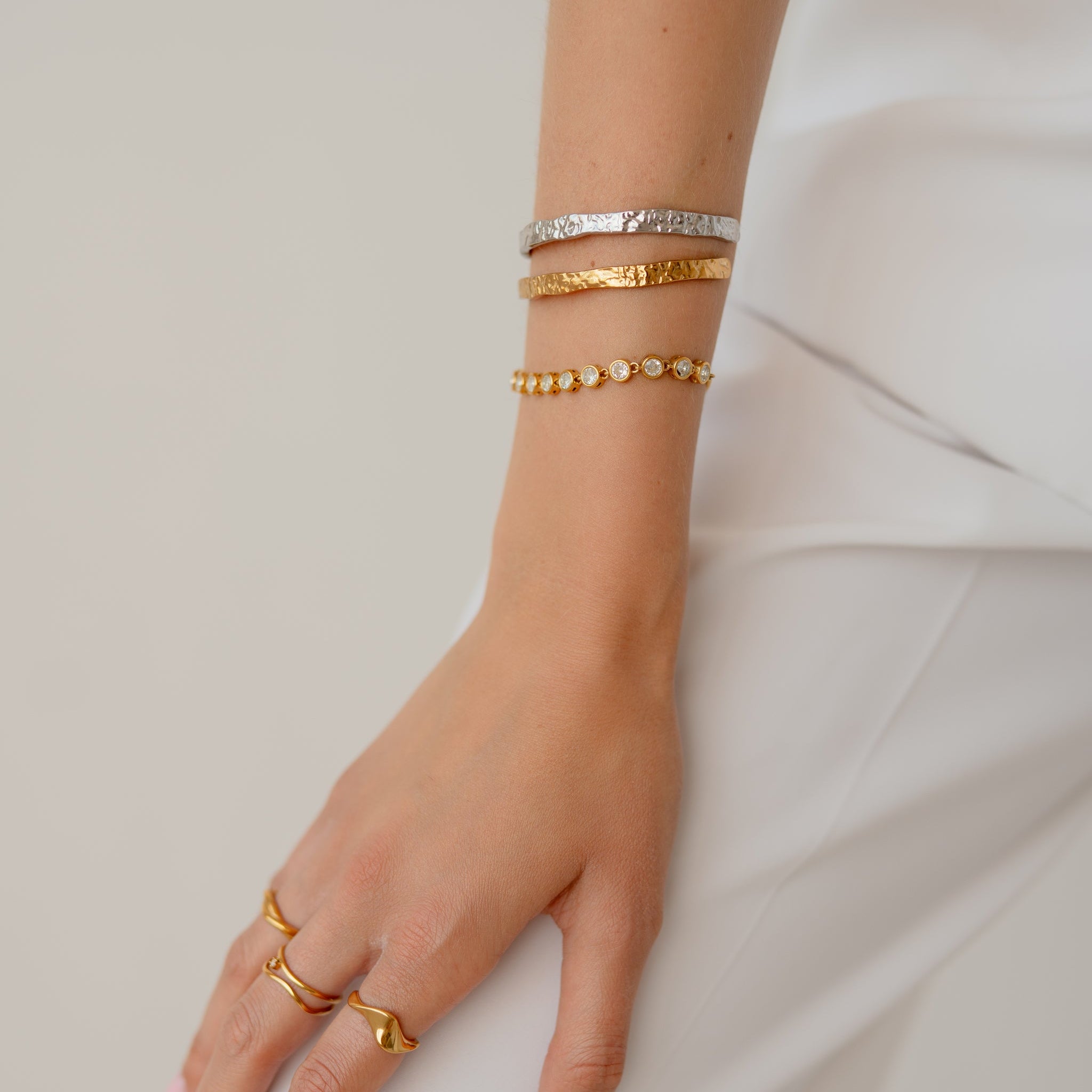 A close-up of a person's hand and wrist wearing multiple delicate bracelets and rings, including gold and silver-toned pieces, against a soft light gray background.