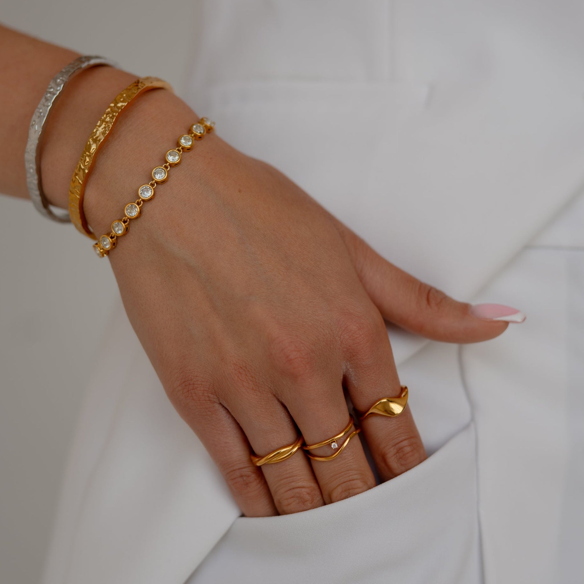 A close-up of a person's hand resting in a white pocket, showcasing several gold bracelets and rings, including a delicate gold beaded bracelet.