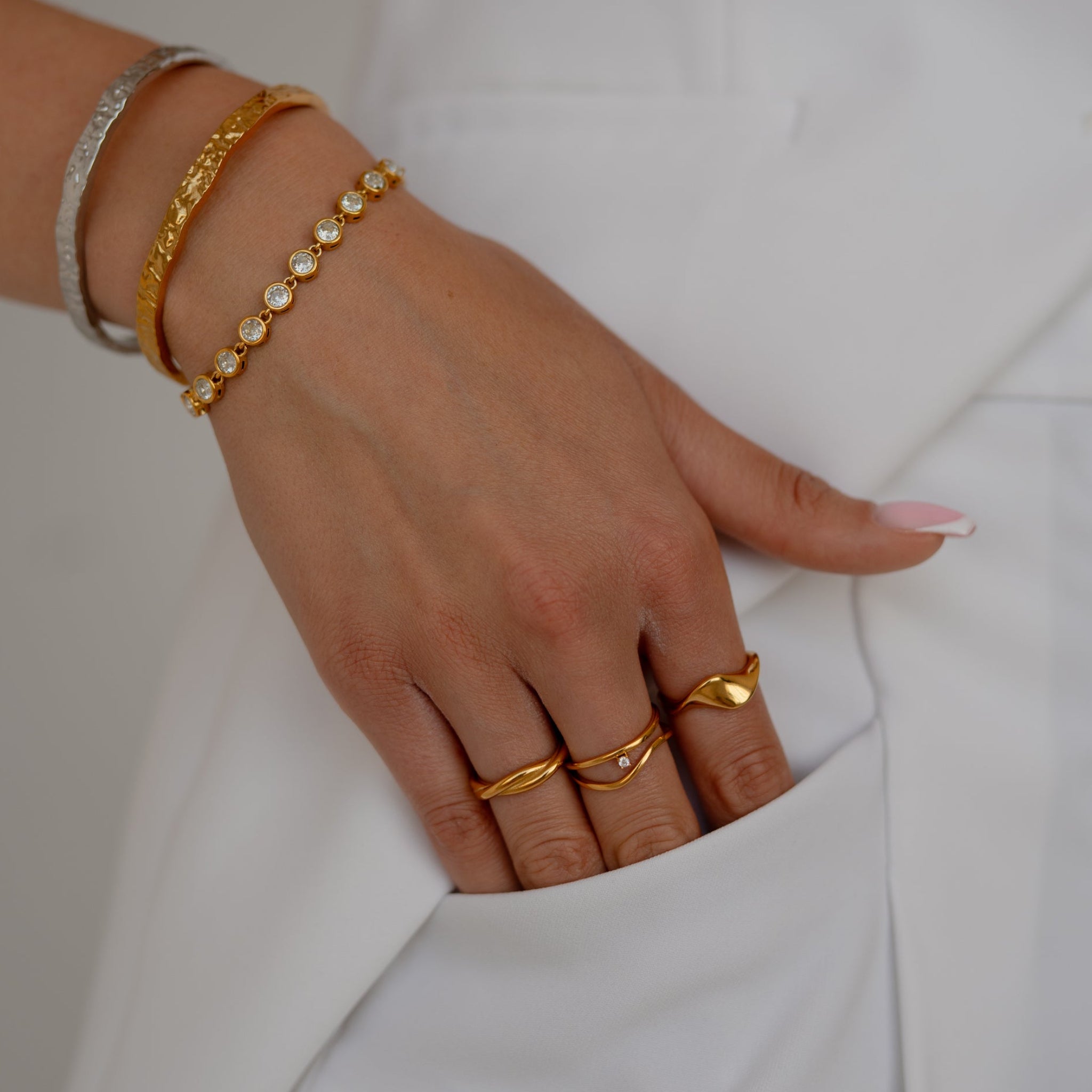 A close-up of a person's hand resting in a white pocket, showcasing several gold bracelets and rings, including a delicate gold beaded bracelet.