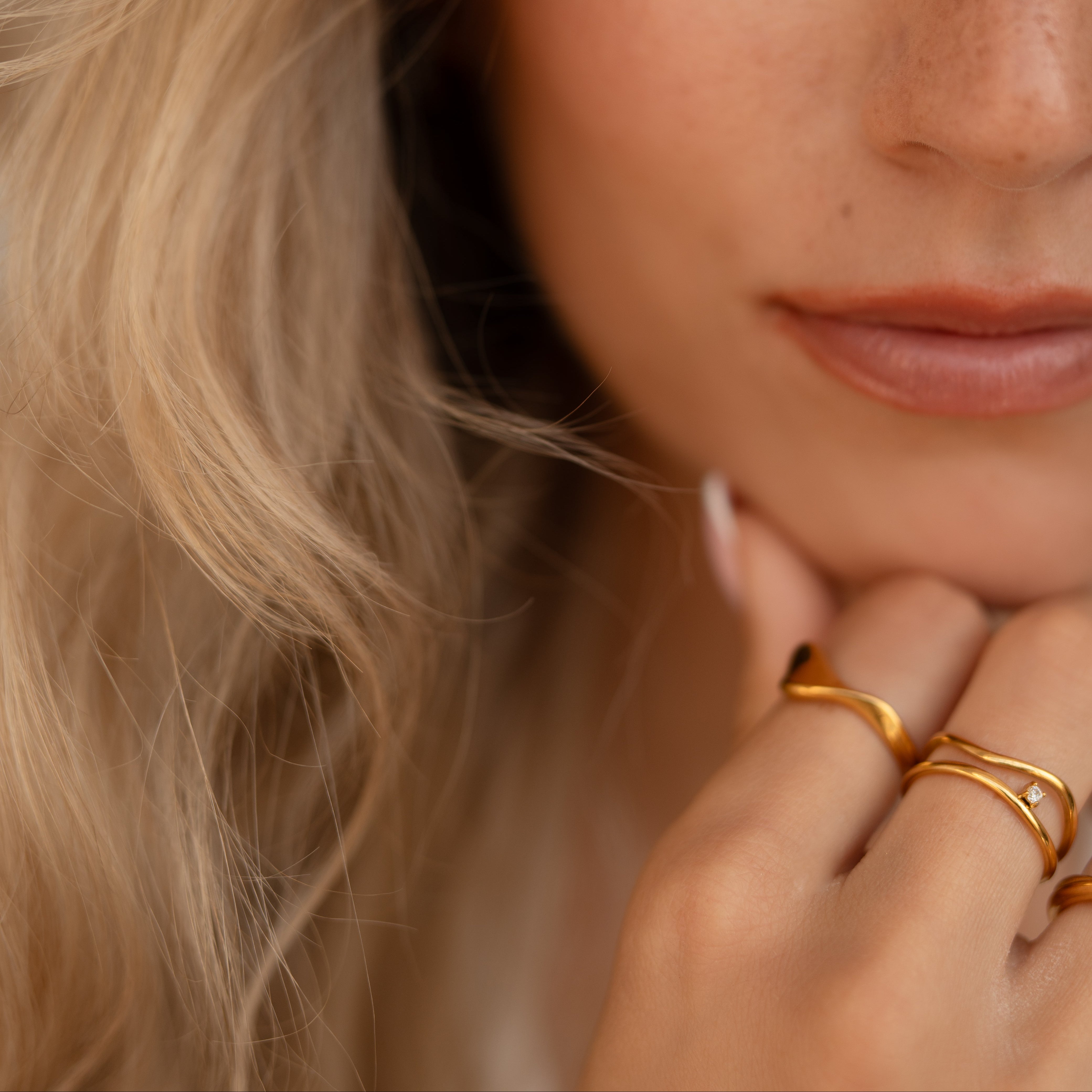 Close-up portrait of a woman with blonde hair resting her chin on her hand, wearing three thin gold rings on her fingers against a soft neutral background.