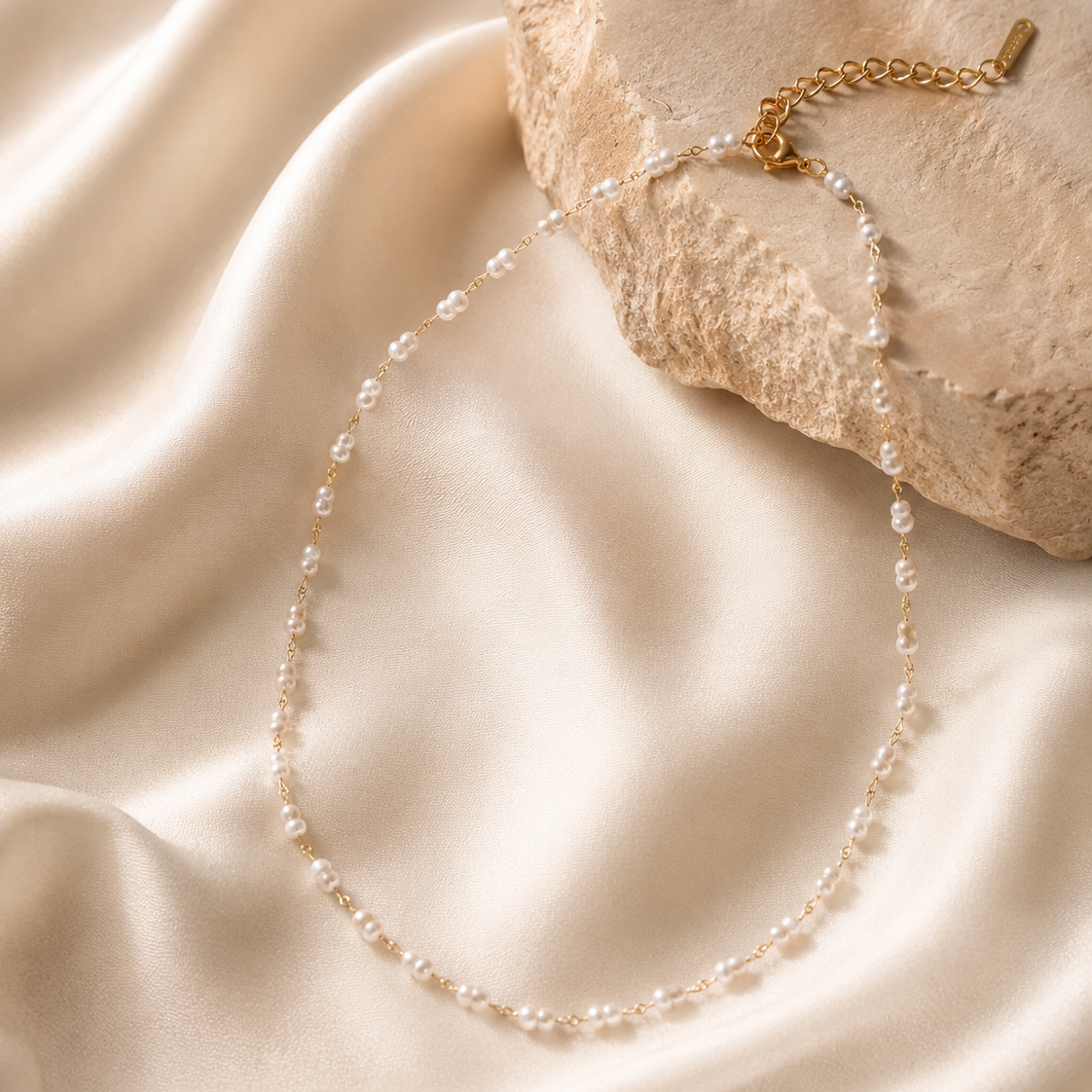 A delicate necklace made of small clear beads on a gold-tone chain, draped over cream satin fabric and a textured stone prop.