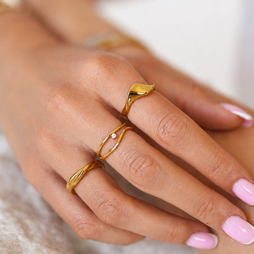A close-up of a person's hand wearing three gold rings, including a leaf-shaped ring and a double-band ring, with light pink manicured nails.