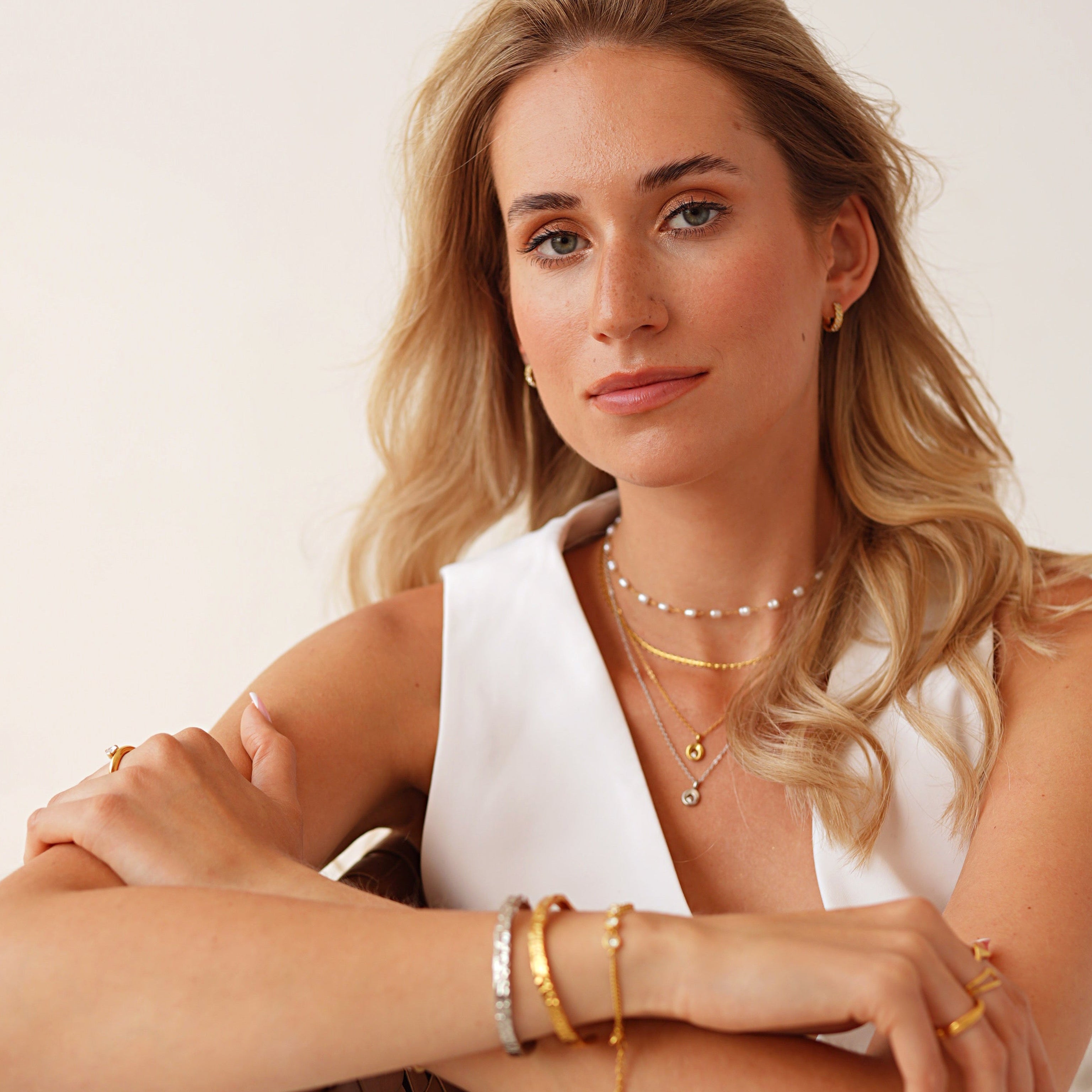A blonde woman wearing layered gold necklaces and bracelets, sitting in a brown woven chair against a plain off-white background.