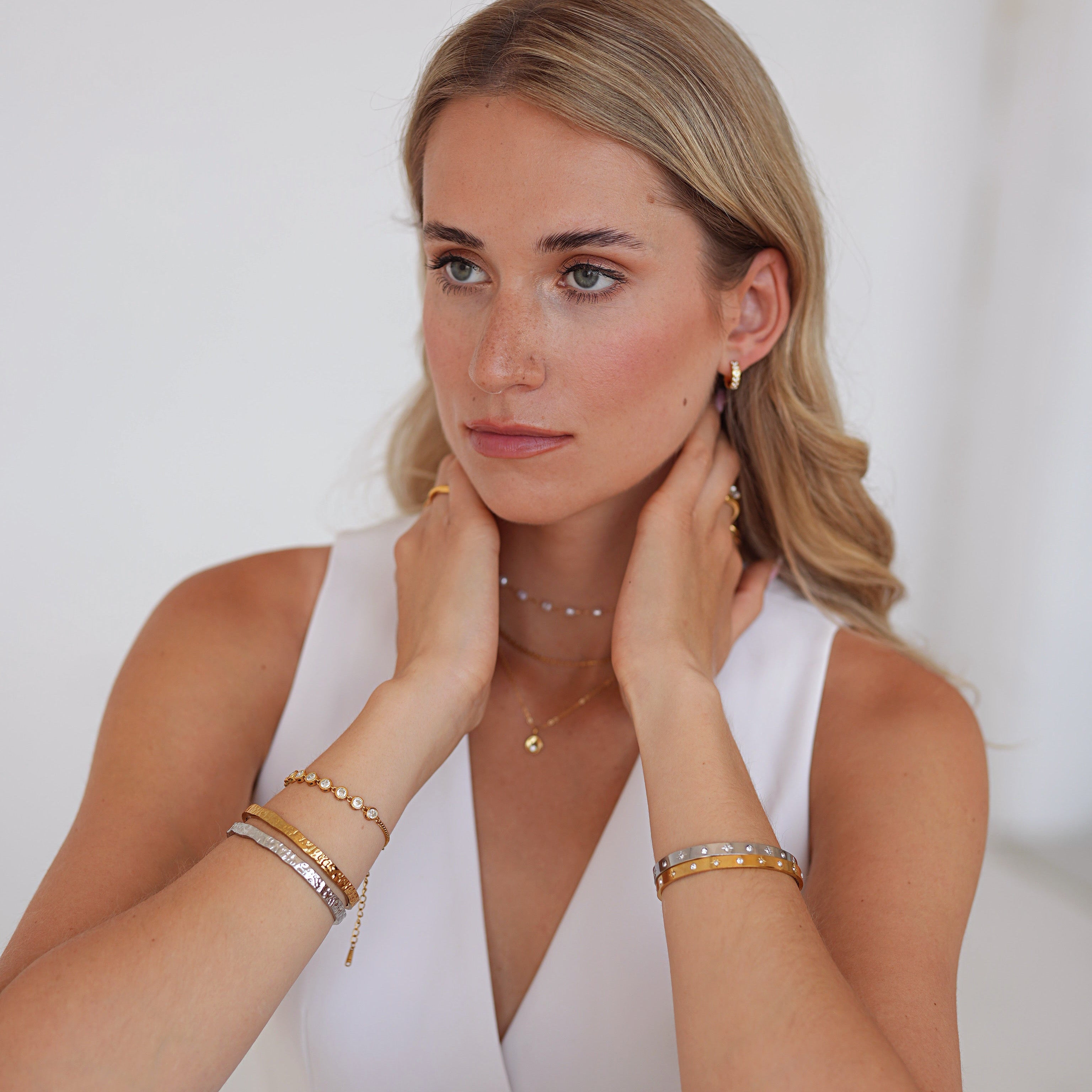 A blonde woman wearing delicate gold jewelry, including stacked bracelets and a necklace, posing in a white sleeveless top against a bright white background.