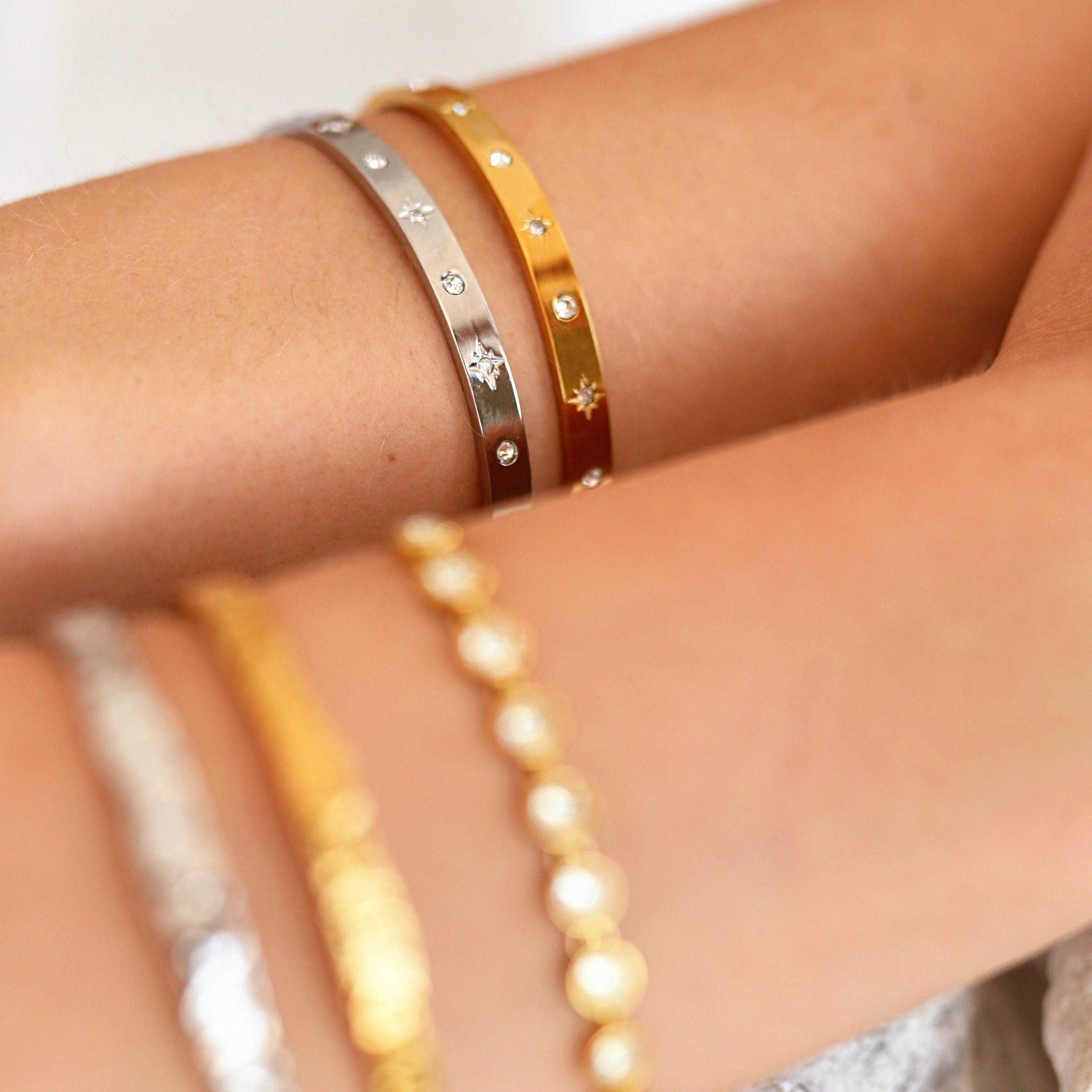 Close-up of a person's wrist wearing multiple delicate bangle bracelets, including gold and silver-toned styles, resting on smooth white stones.