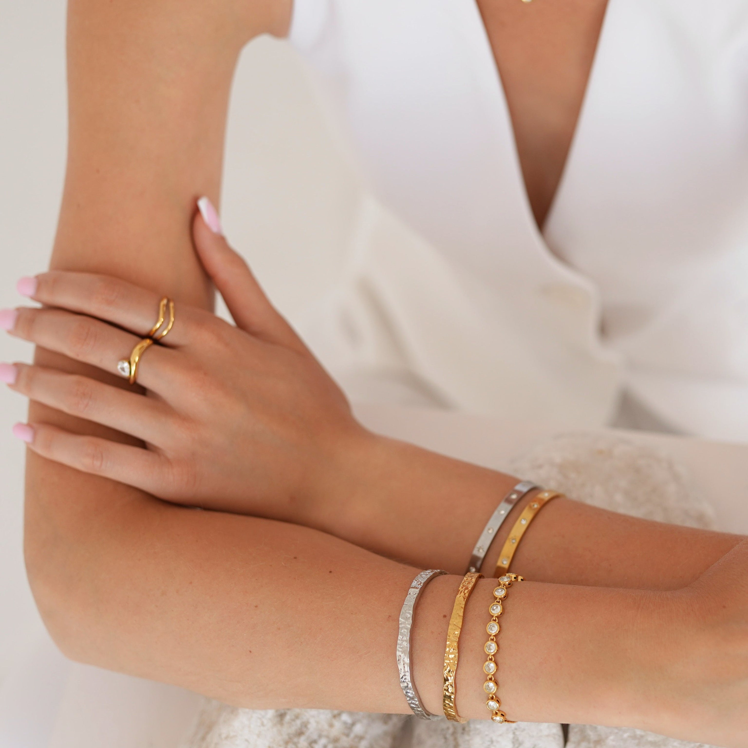 A close-up of a person wearing multiple delicate gold bracelets and rings on both wrists and fingers, paired with a white sleeveless top against a soft light background.