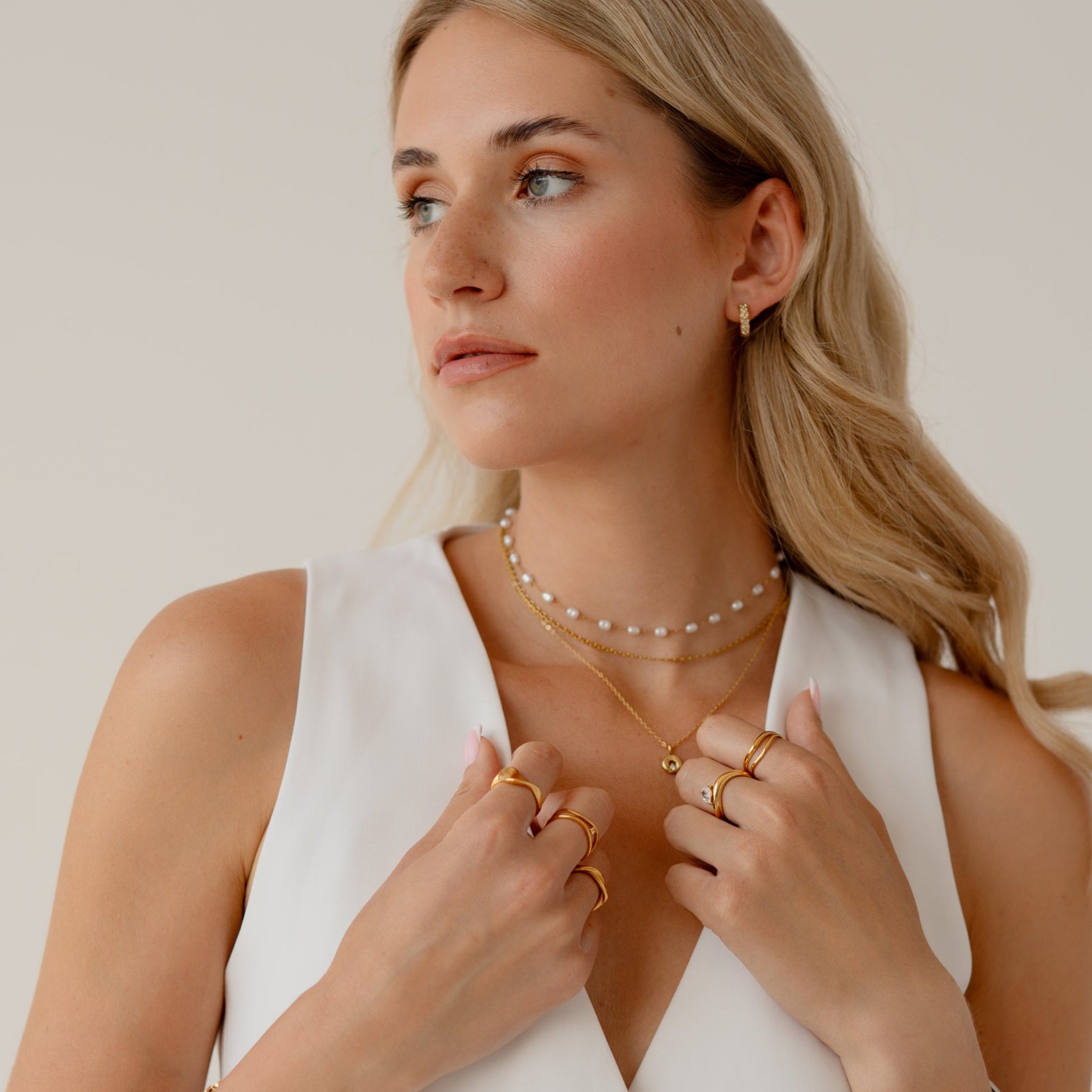 A blonde woman wearing a white sleeveless vest and multiple delicate gold and pearl necklaces, posing against a light beige studio background.