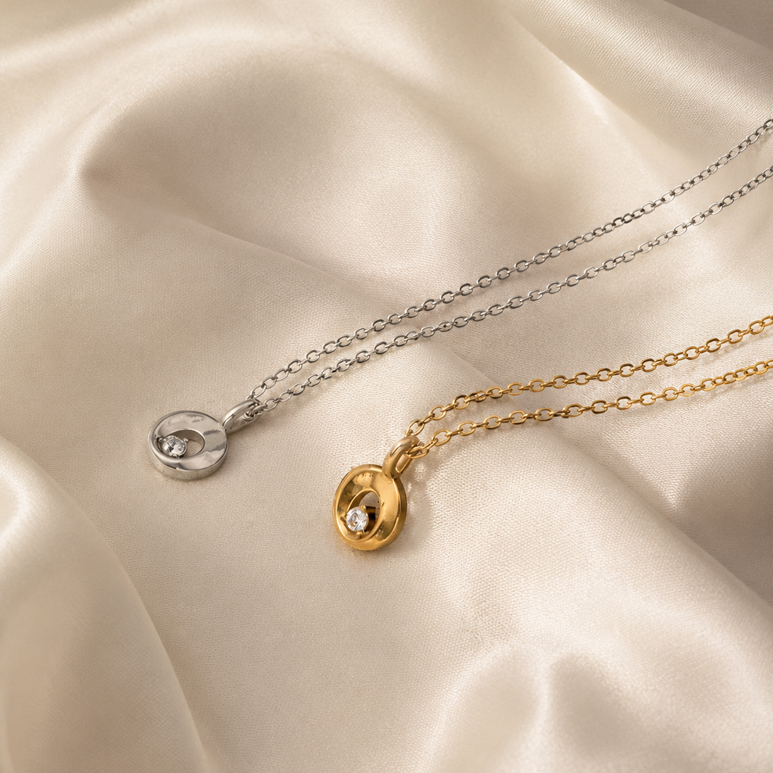 Two delicate pendant necklaces laid on draped satin fabric: one silver-toned chain with a small oval silver pendant and one gold-toned chain with a small oval gold pendant, each with a clear stone.