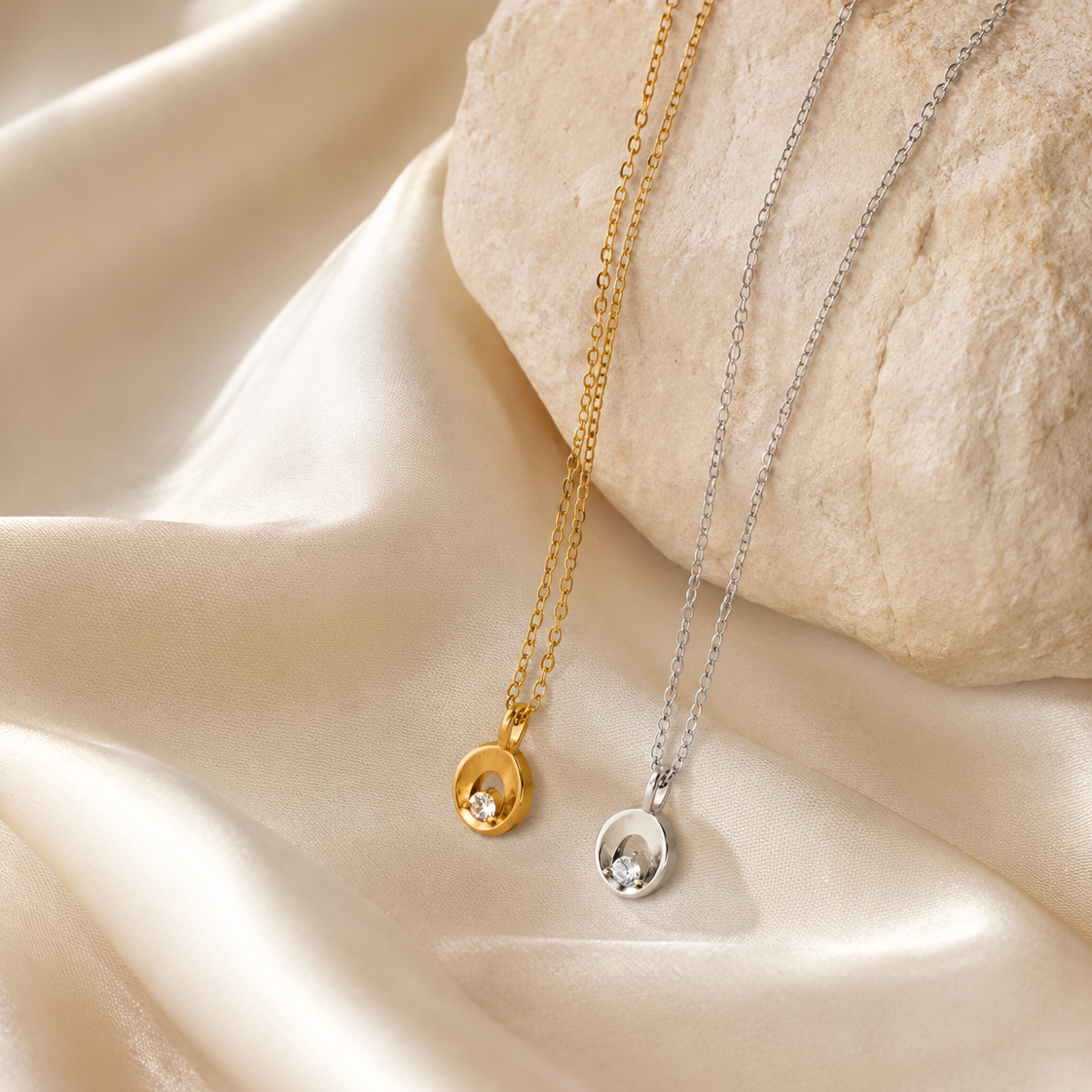 Two delicate pendant necklaces draped over a smooth stone on draped satin fabric. One necklace is gold-toned with a small oval pendant holding a clear stone, and the other is silver-toned with a matching oval pendant.