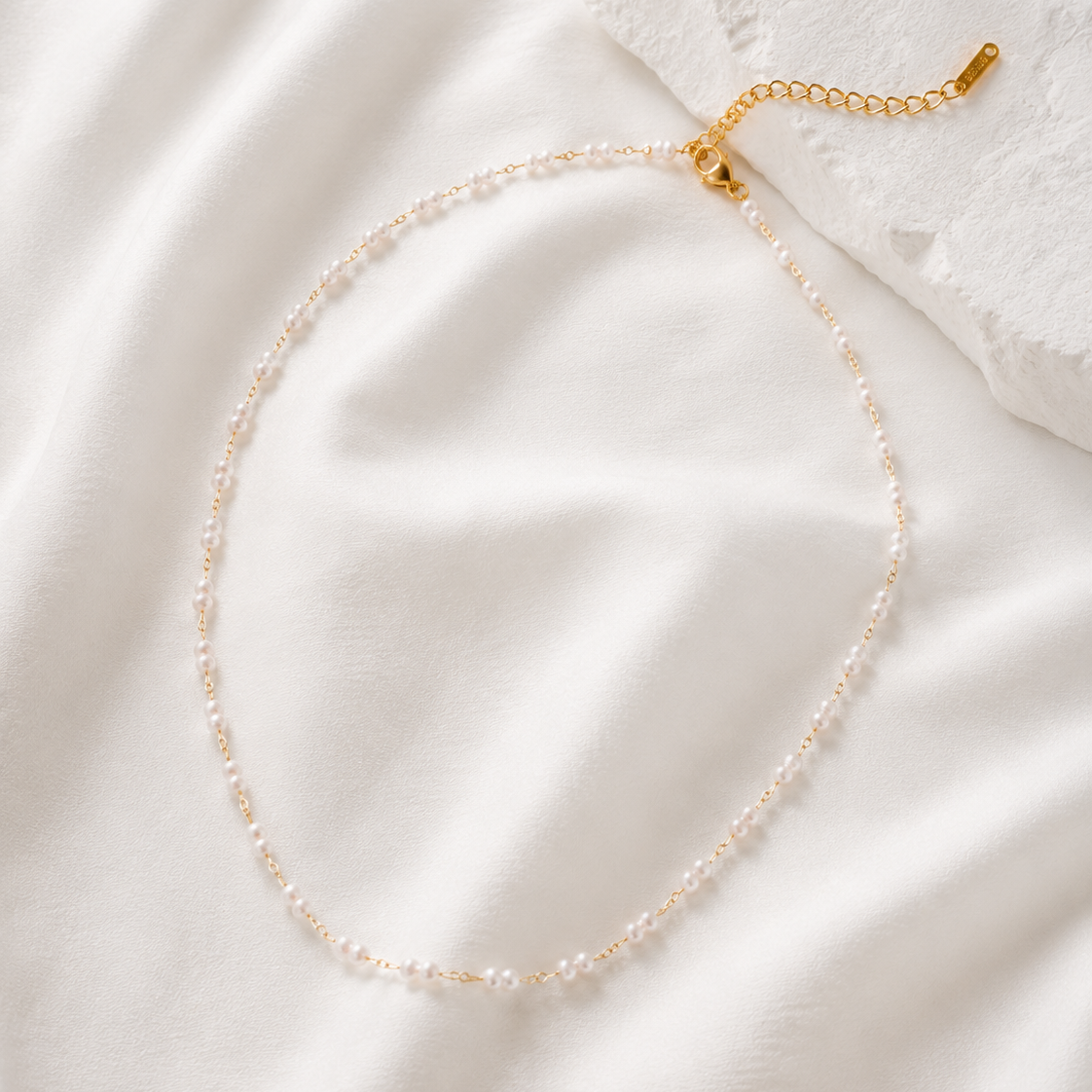 A delicate necklace made of small clear beads on a gold chain, arranged in an oval on white draped fabric with a textured white stone prop.