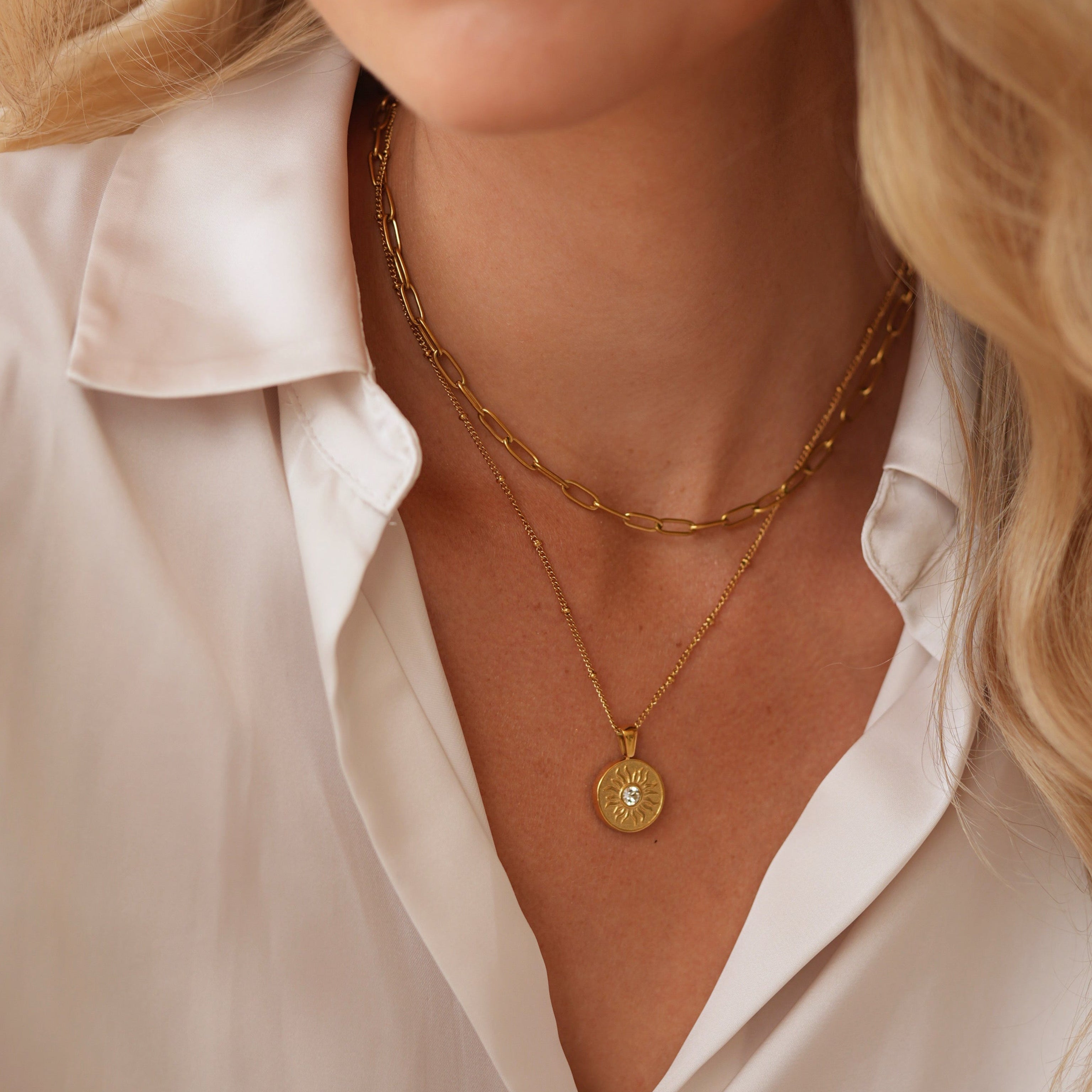A close-up of a blonde woman wearing layered gold necklaces, including a delicate chain necklace and a longer chain with a round gold pendant, paired with a cream satin button-up shirt.