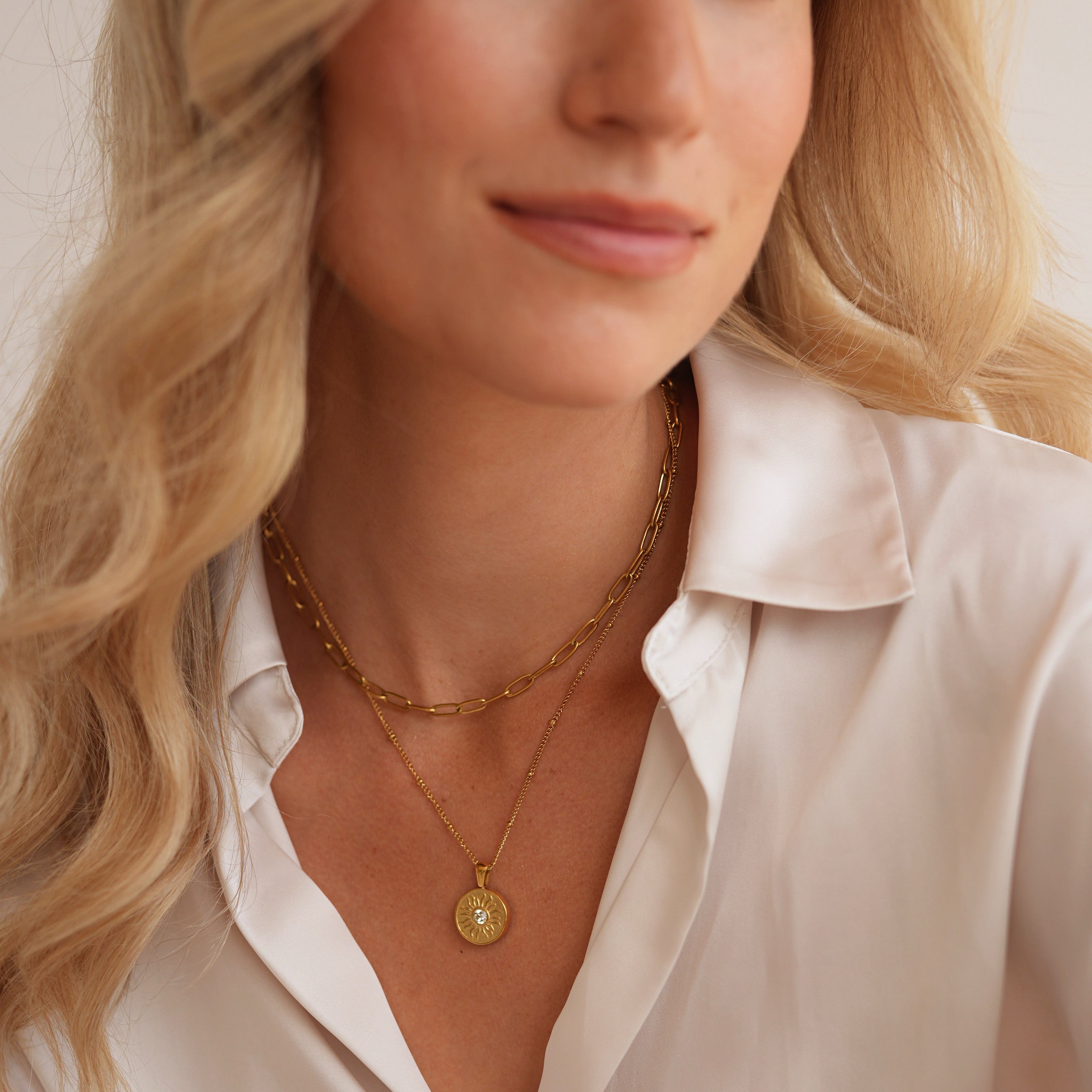 A close-up of a blonde woman wearing layered gold necklaces, including a delicate chain necklace and a longer chain with a round gold pendant, paired with a cream satin button-up shirt.