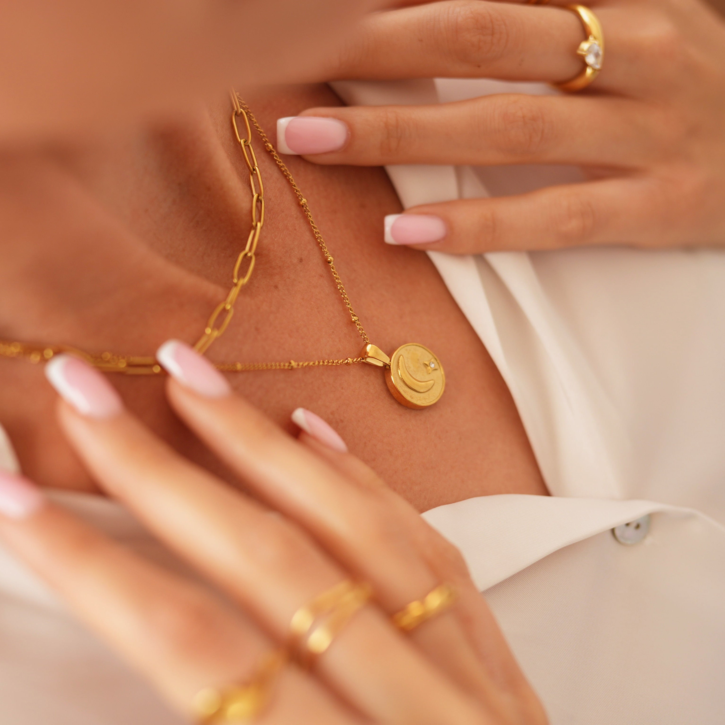 Close-up of a person wearing layered gold necklaces and rings, with manicured pink nails and a white collared shirt visible.